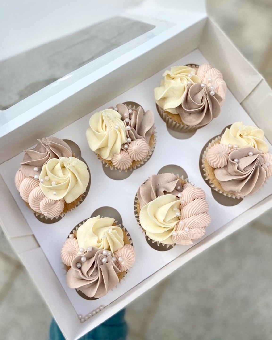 A person is holding a box of cupcakes with pink and white frosting
