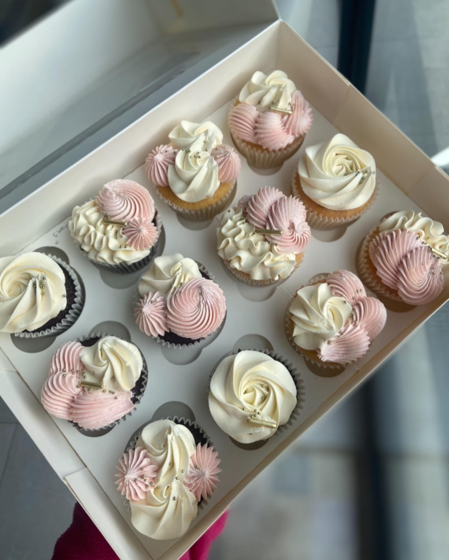 A person is holding a box of cupcakes with pink frosting