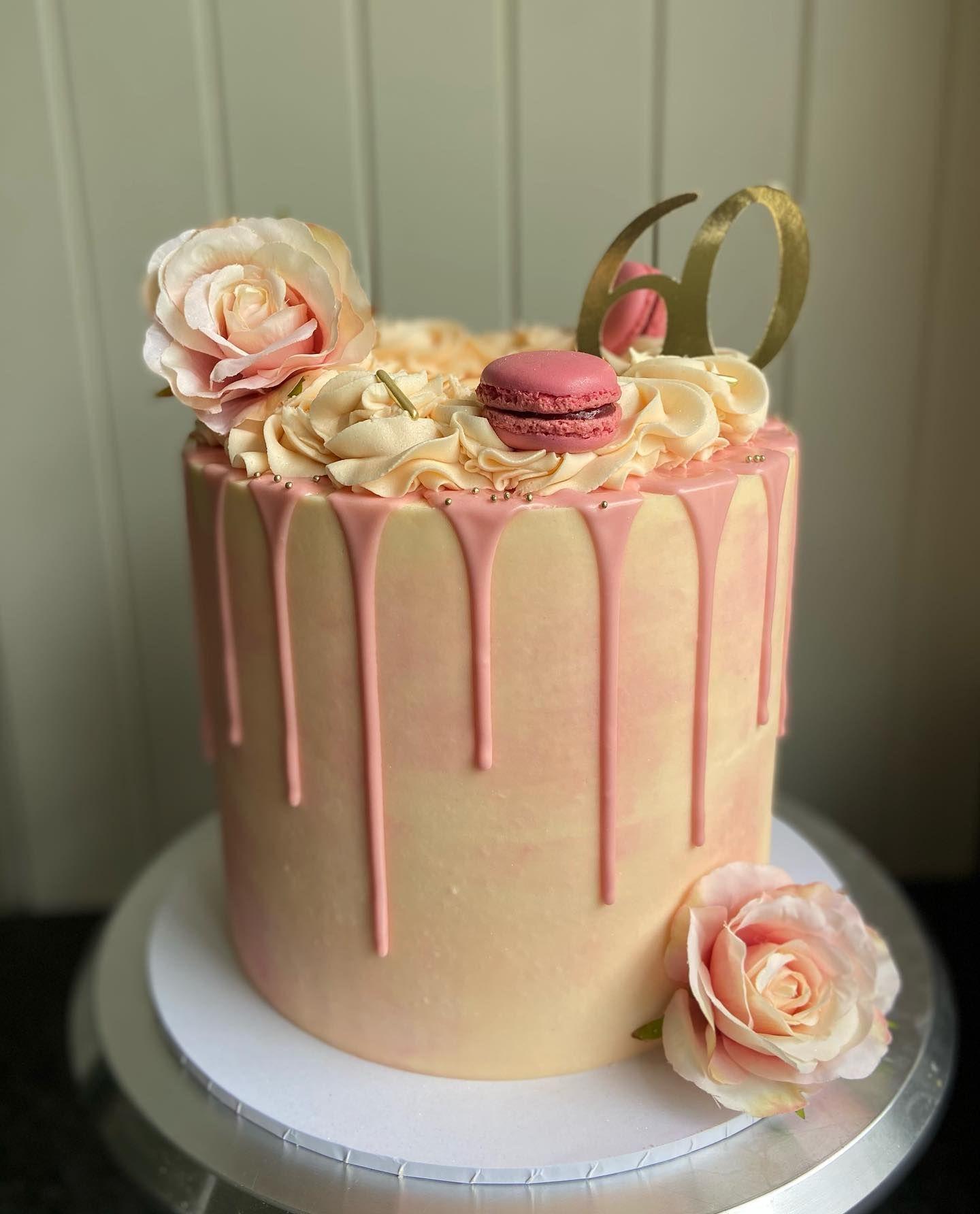 A pink drip cake with roses and macarons on top is on a cake stand.