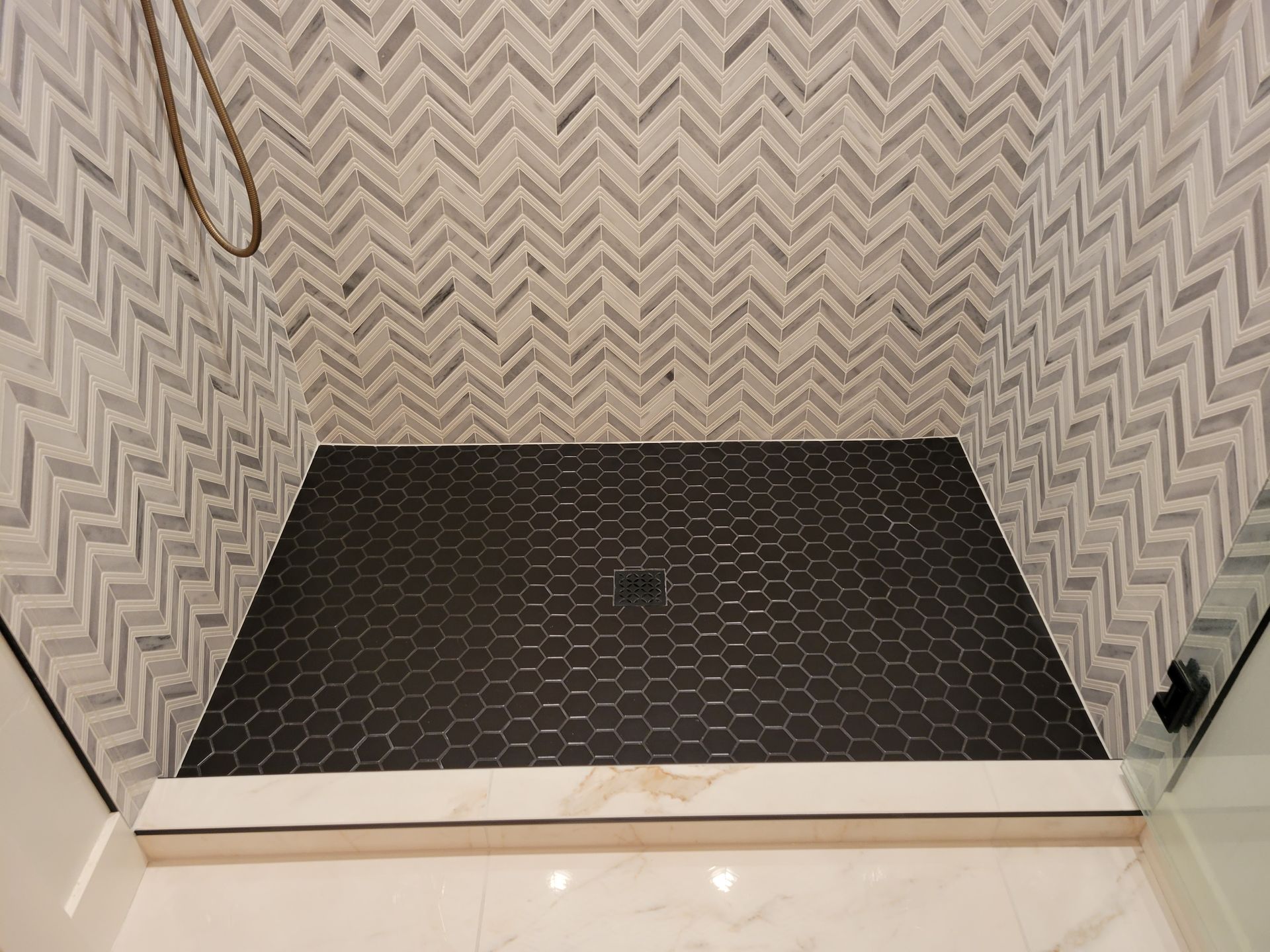 A walk in shower with a black tile floor and a herringbone pattern on the wall.