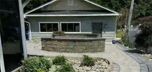 A light blue building with windows and a gray stone wall in front of it. A brick path leads to the building.