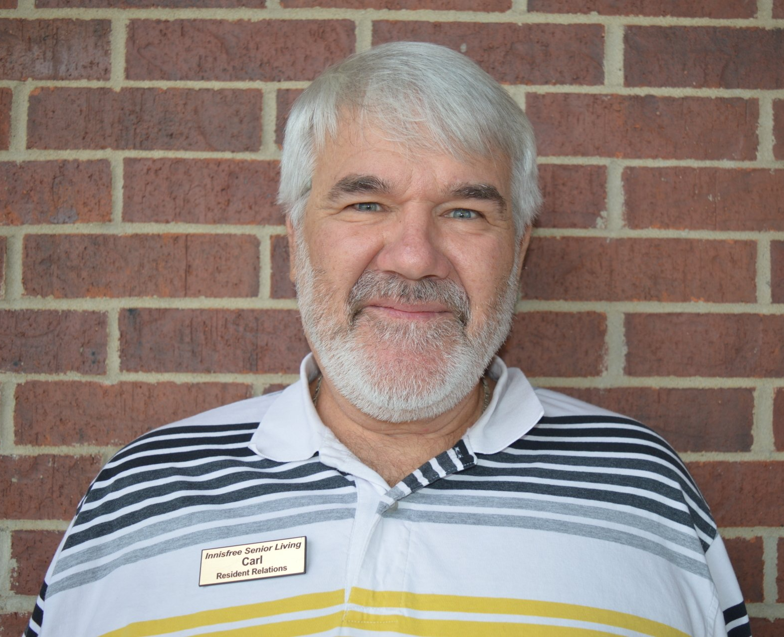 Man with gray hair and beard, wearing a striped polo shirt, standing in front of a brick wall.