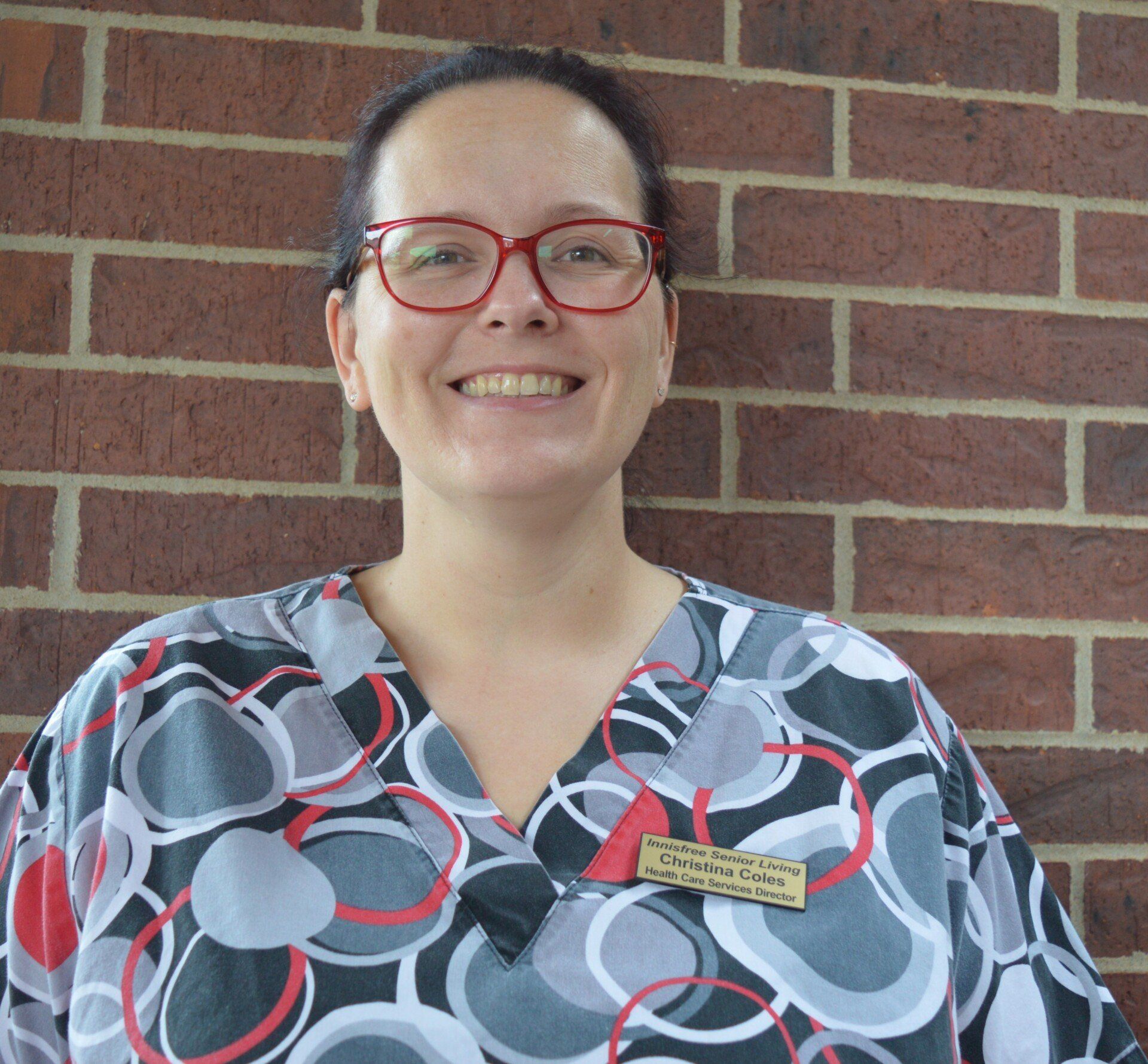 Woman in glasses, patterned scrubs smiles against a brick wall. Name tag visible.