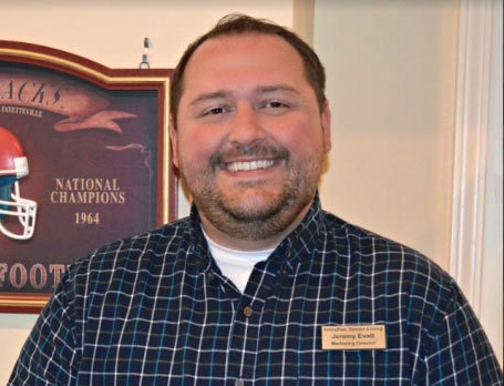 Man with a name tag smiling, in a blue plaid shirt, in front of a sports memorabilia plaque.