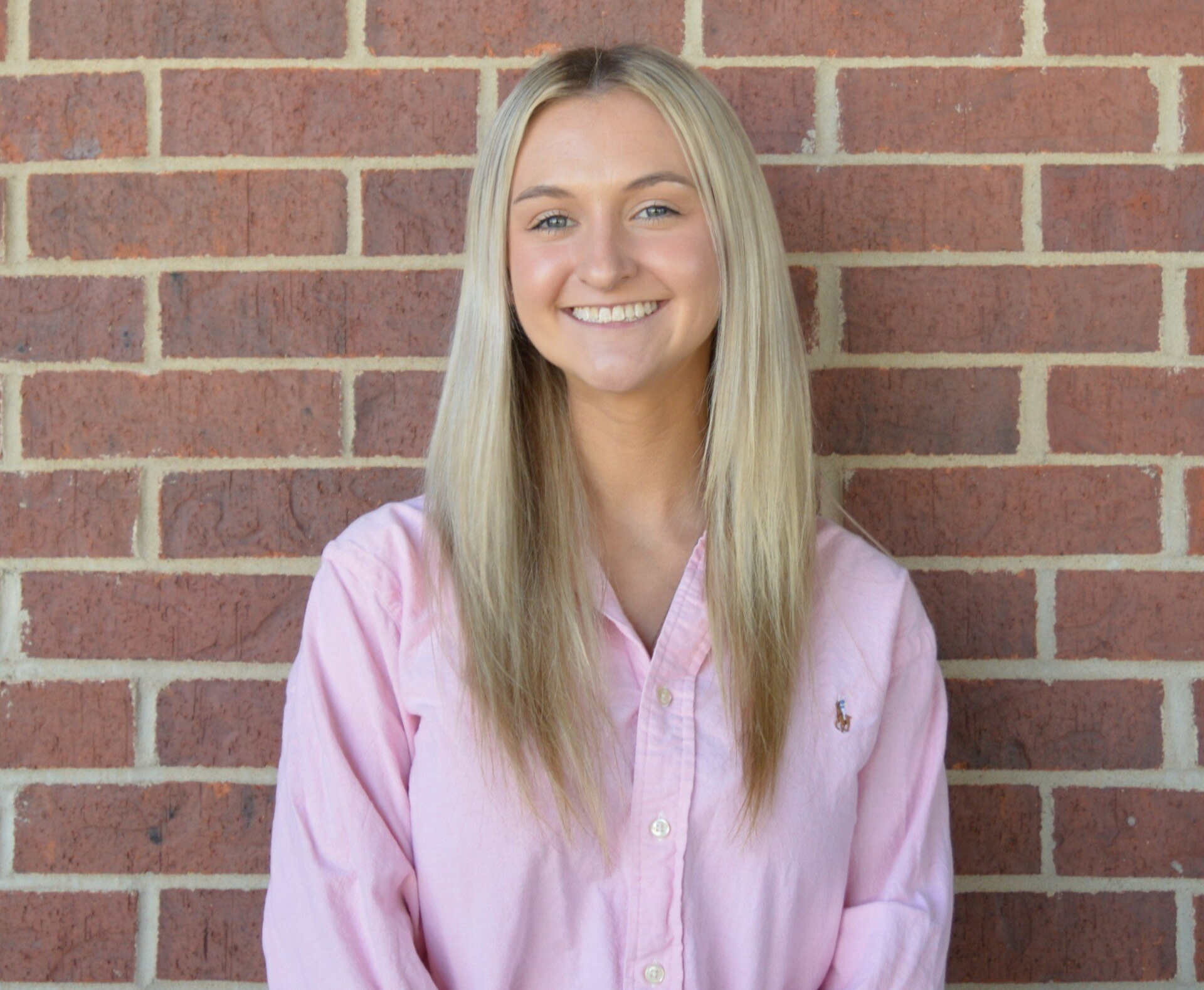 Blonde woman in pink shirt smiles in front of a brick wall.