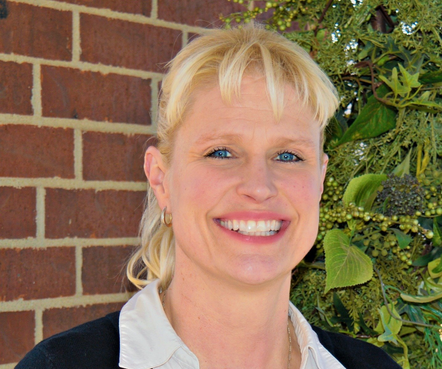 Blonde woman smiling, wearing a black sweater over a light shirt; name tag visible.