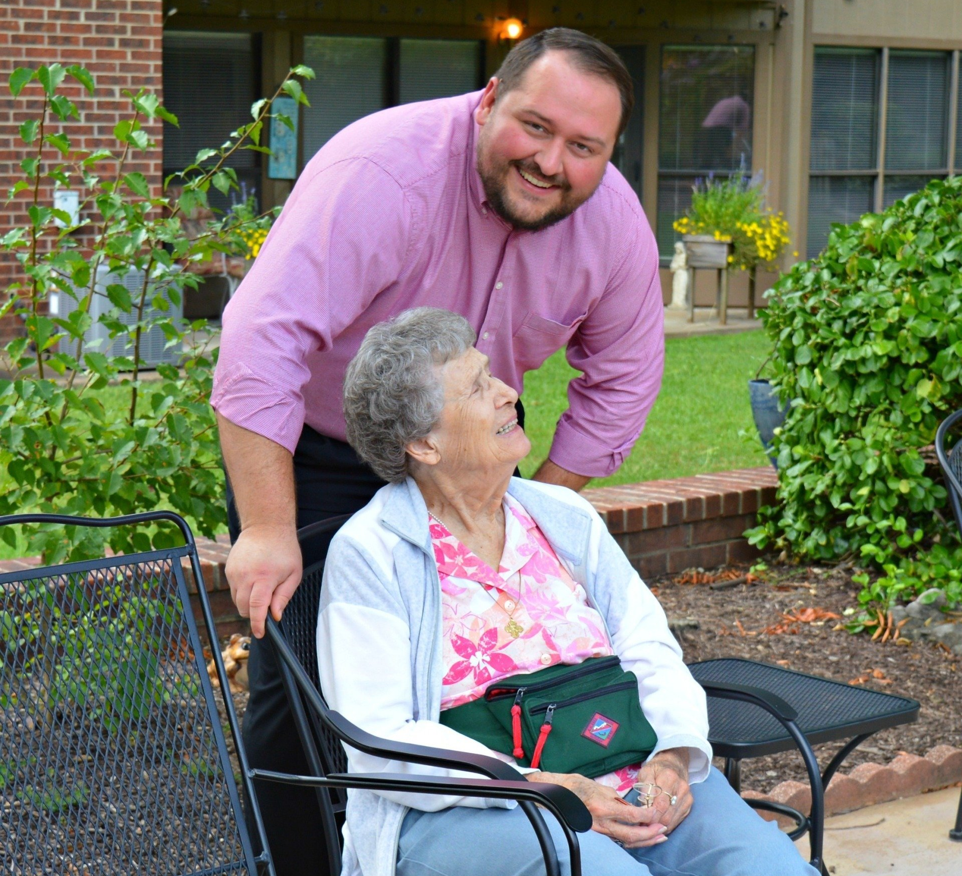 Man smiles, leans over woman seated outside. Man wears pink shirt, woman in light jacket, smiling.