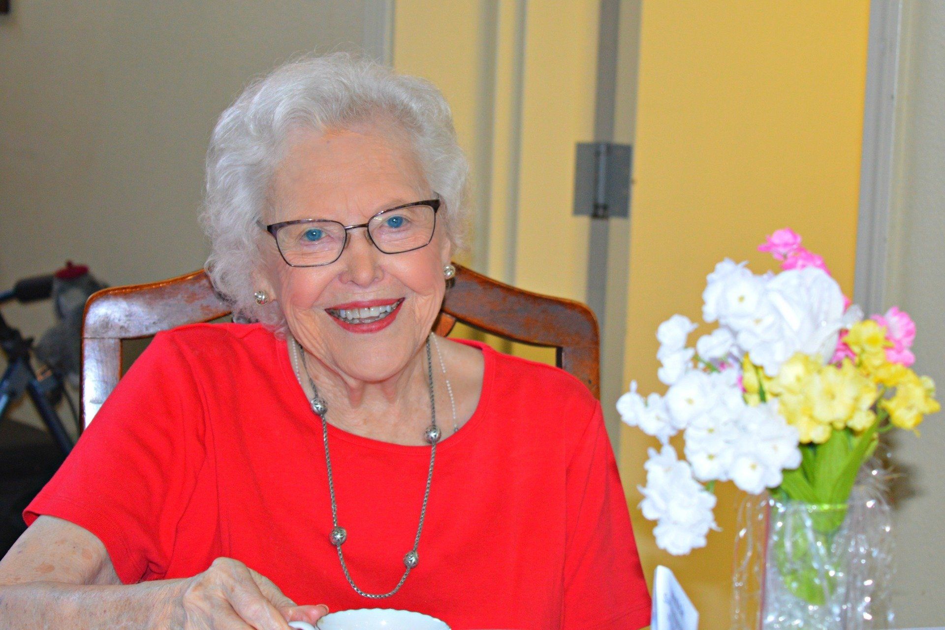 Smiling woman wearing glasses and red shirt, sitting at a table with flowers.