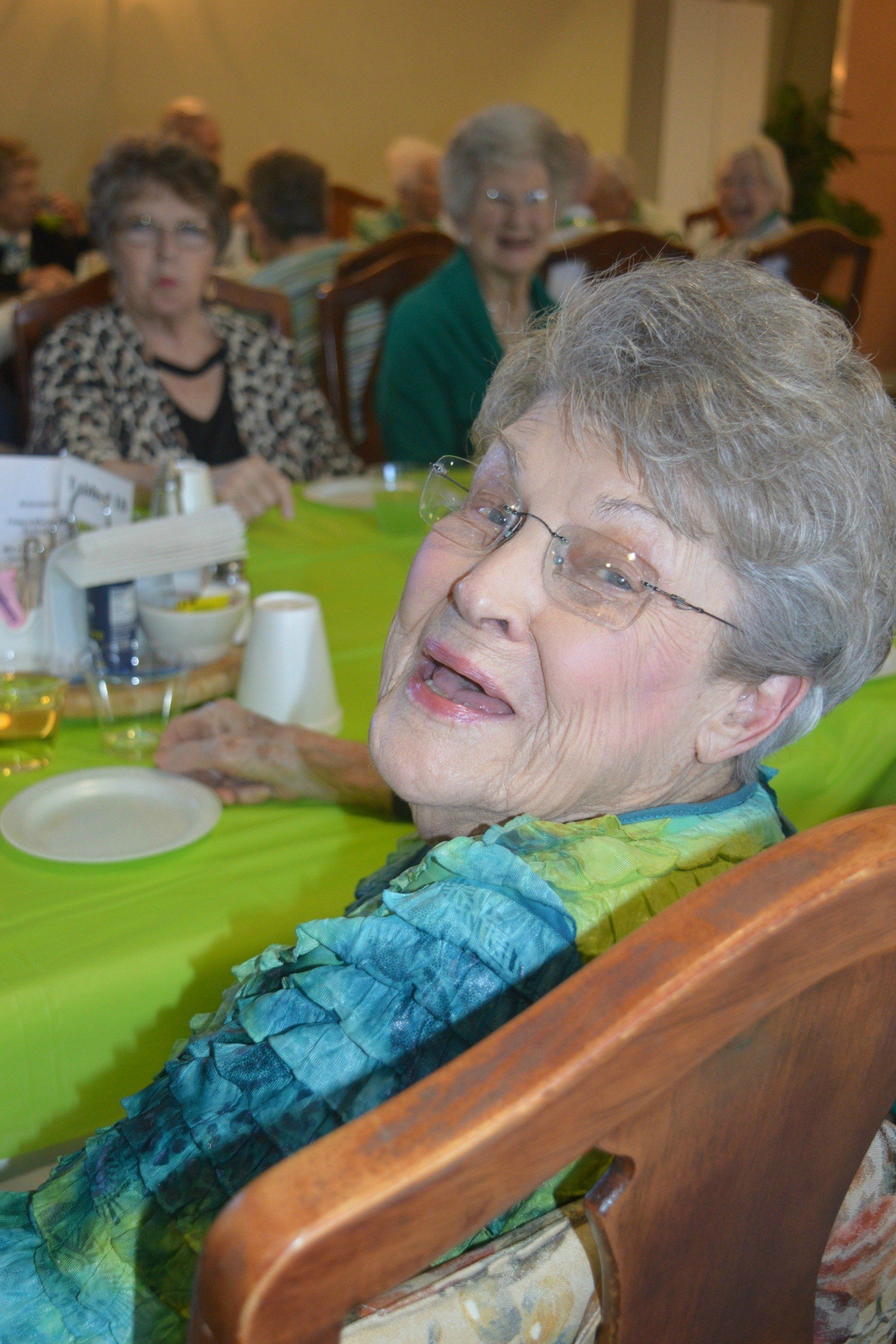 Woman smiling at a table, possibly at an event, with other people and green tablecloth.