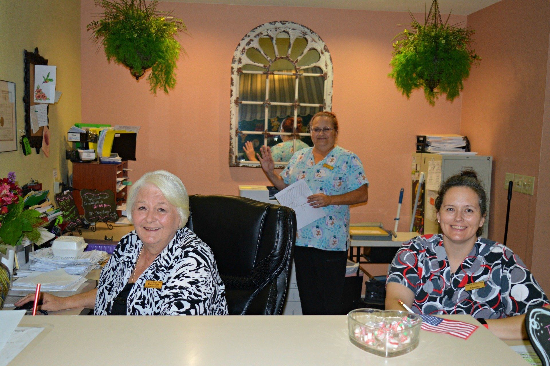 Three people at a reception desk in an office. One waves, another smiles, the third sits.