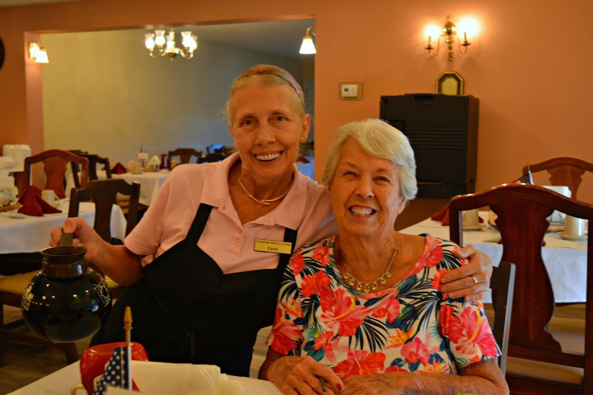 Woman in uniform and older woman smiling in a dining room; the woman in uniform holding a pitcher.