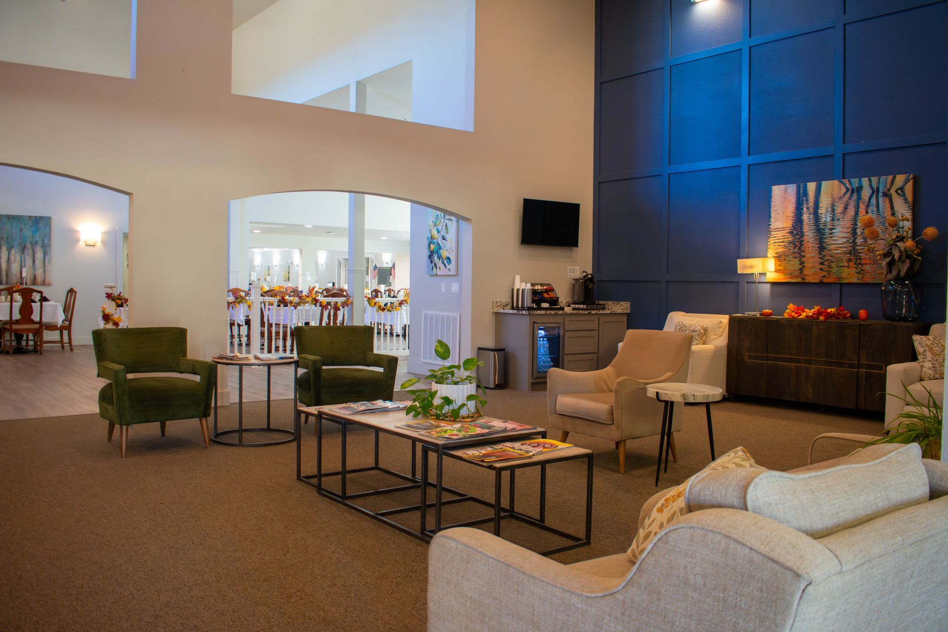 Lounge area with seating, tables, and art. A doorway leads to a dining hall. Blue wall and neutral color scheme.