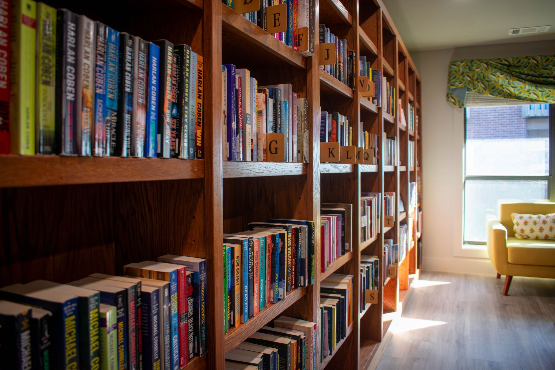 Wooden bookshelf filled with colorful books, in a room with a yellow chair and a window.