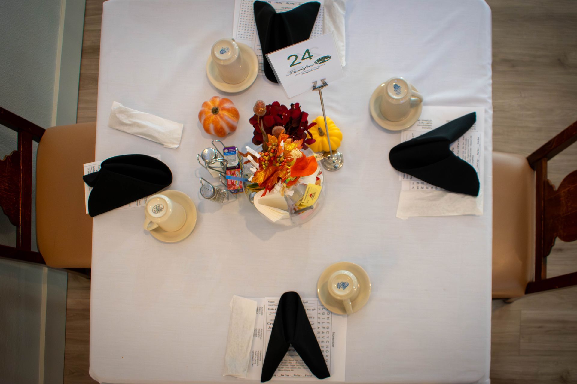 Overhead view of a dining table set for five people. Centerpiece of pumpkins and flowers. Beige teacups. Black napkins.