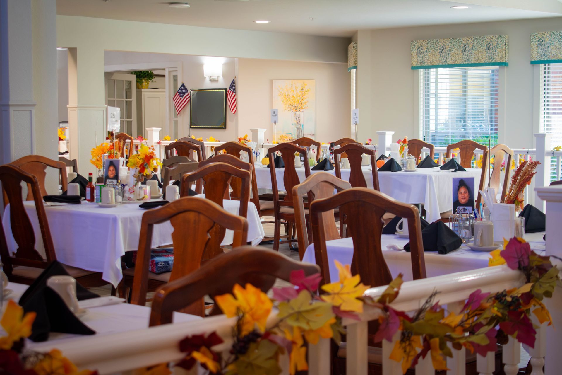 Dining room set for a celebration. Tables covered in white tablecloths, decorated with fall foliage.