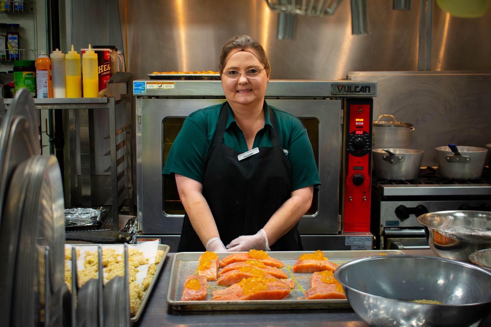 Woman in a kitchen apron and gloves, standing behind a tray of seasoned salmon fillets.