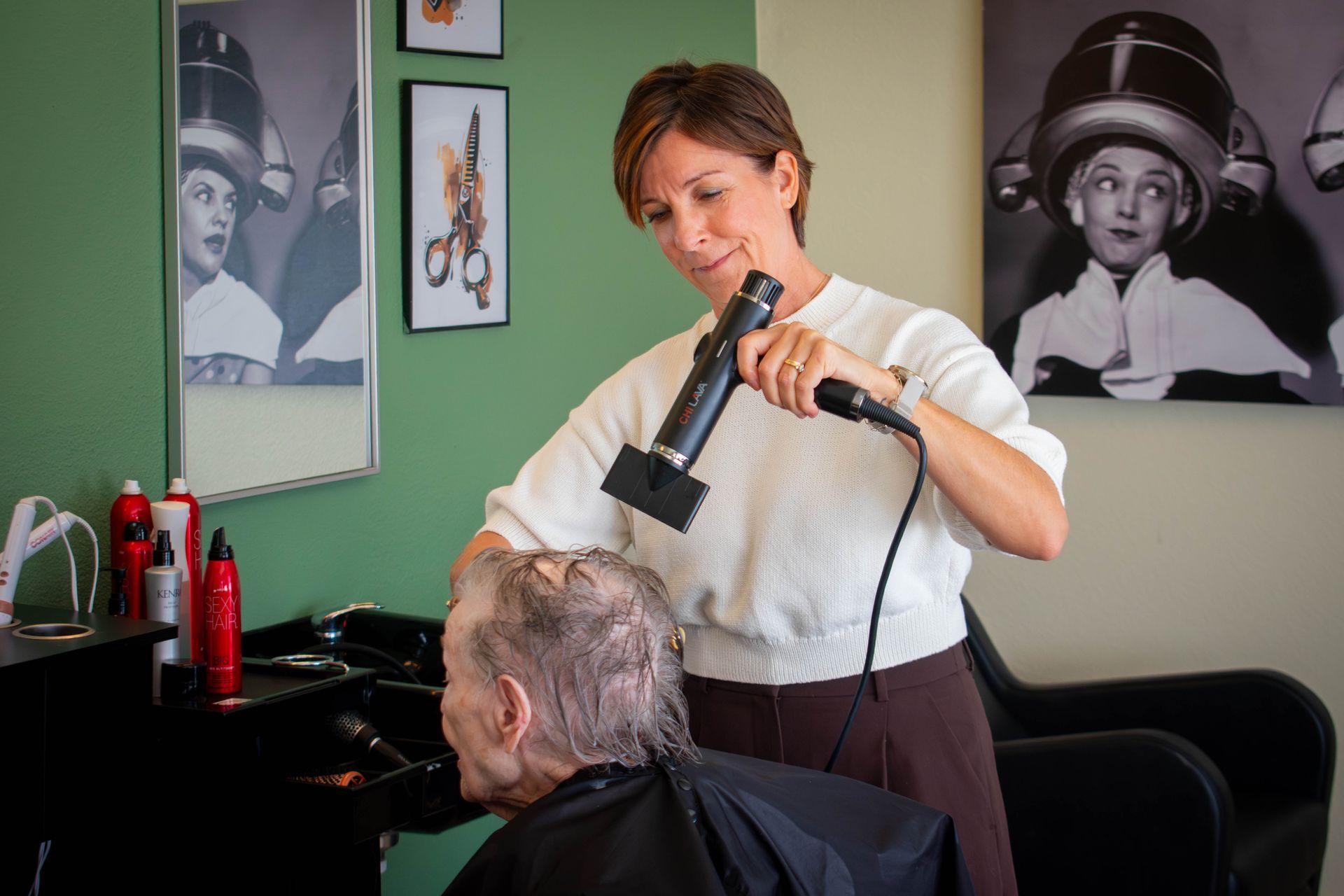 Hairstylist blow-drying client's hair at a salon. Green wall, vintage photos in background.