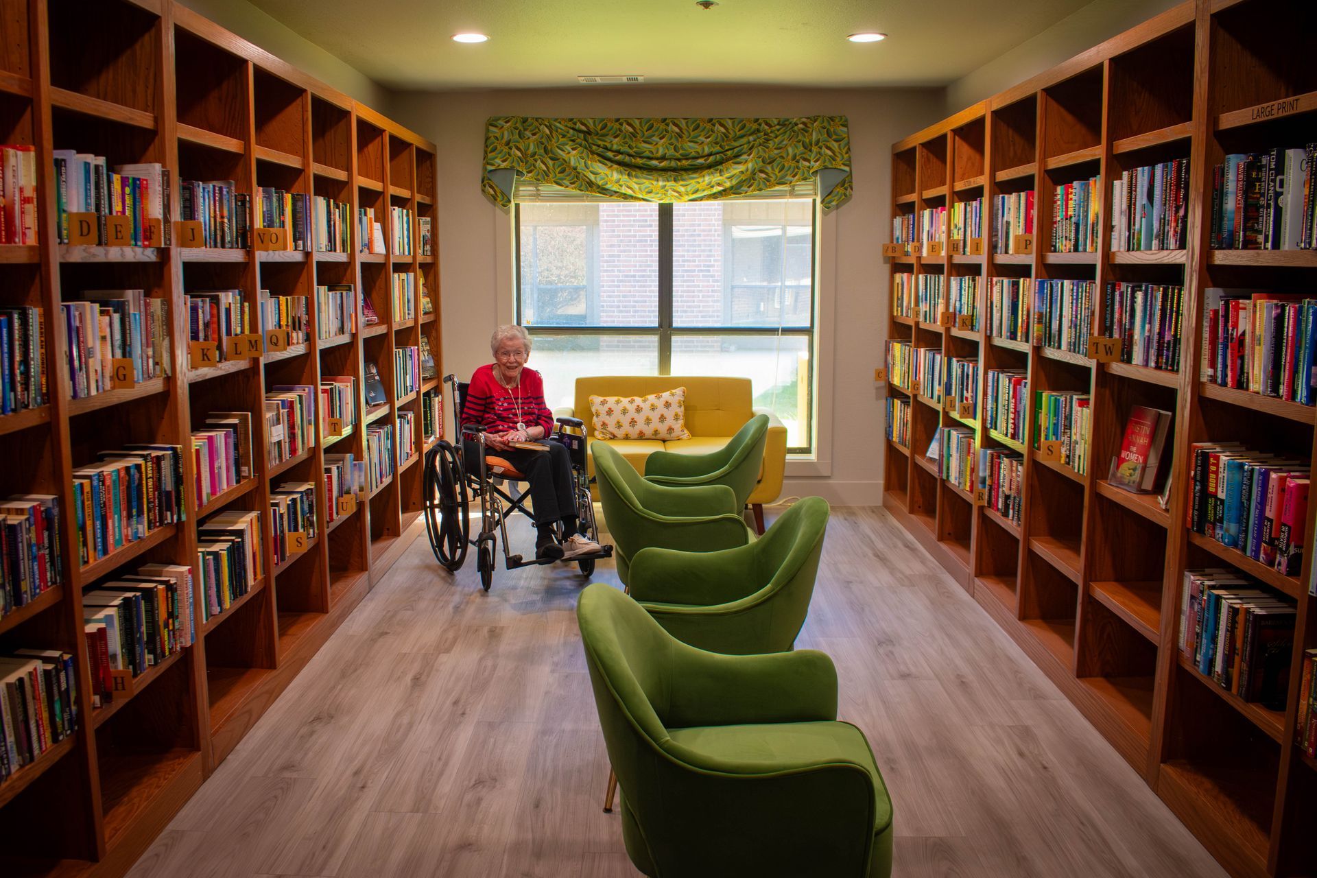 Library interior with bookshelves, a person in a wheelchair, and green chairs.