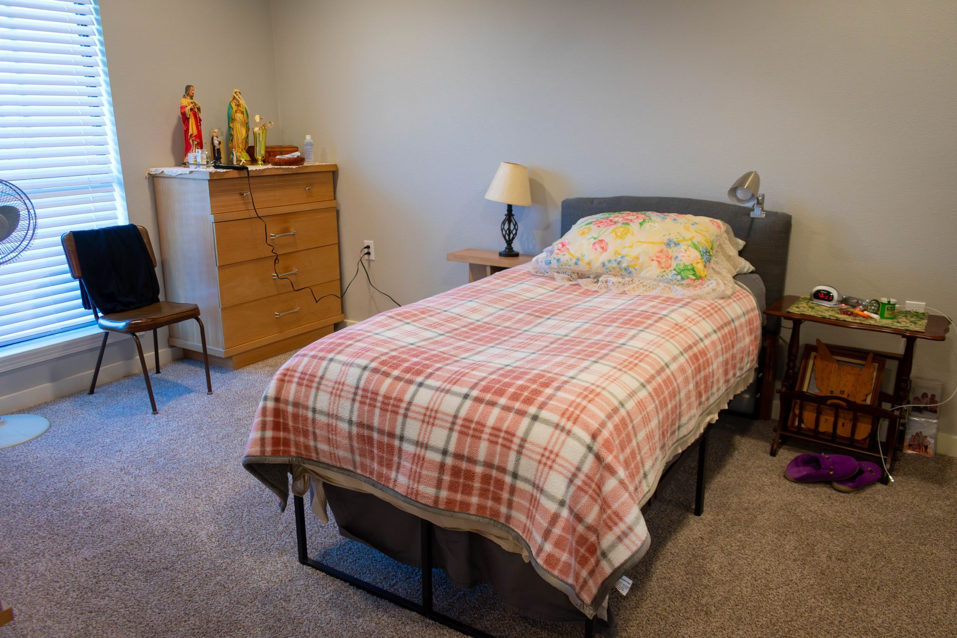 Bedroom with bed, dresser, chair, window, lamp, and side table.  Red and white plaid blanket. Gray walls and carpet.