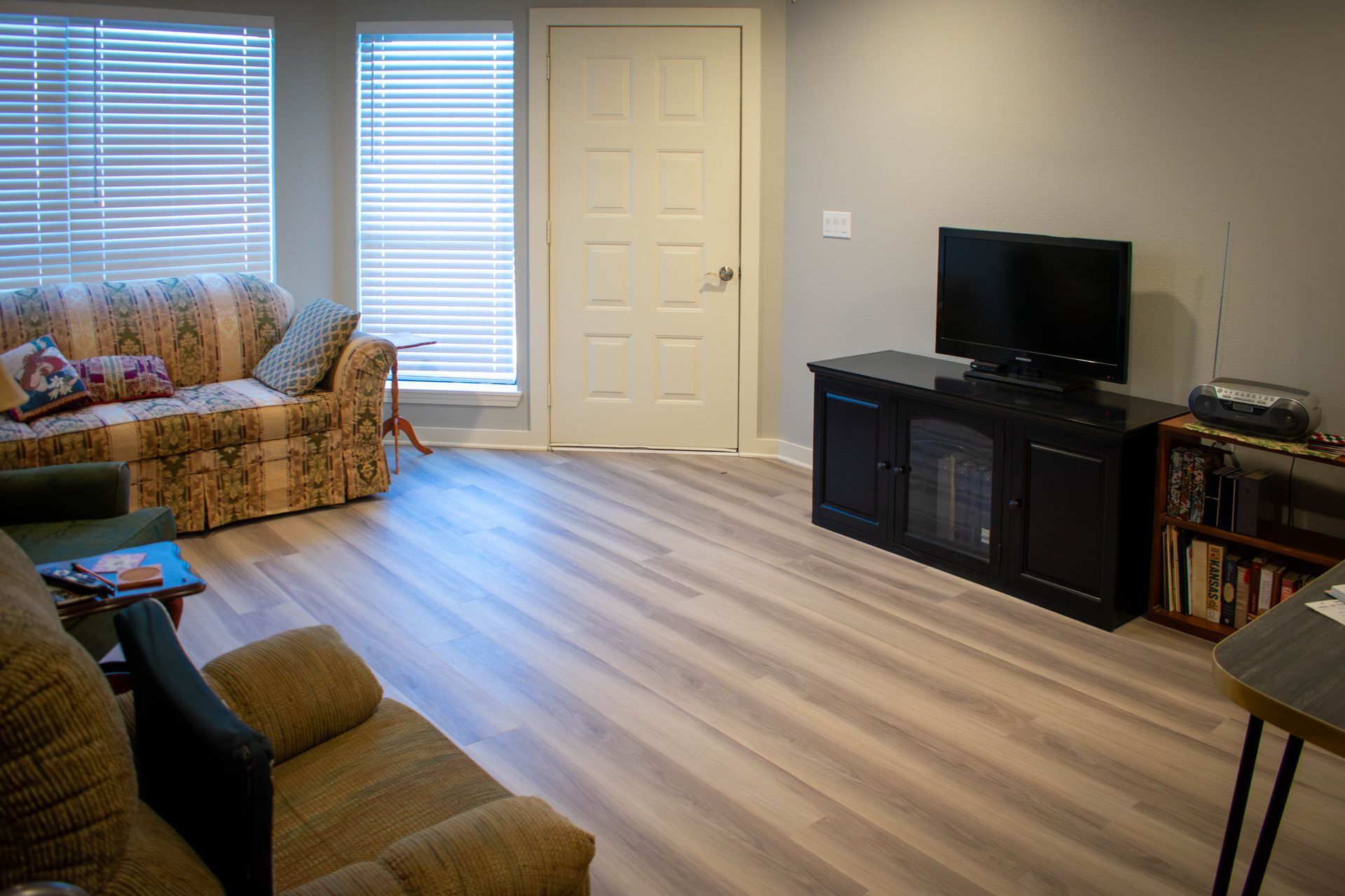 Living room with couches, a TV on a black cabinet, and a door. Natural light comes through blinds.