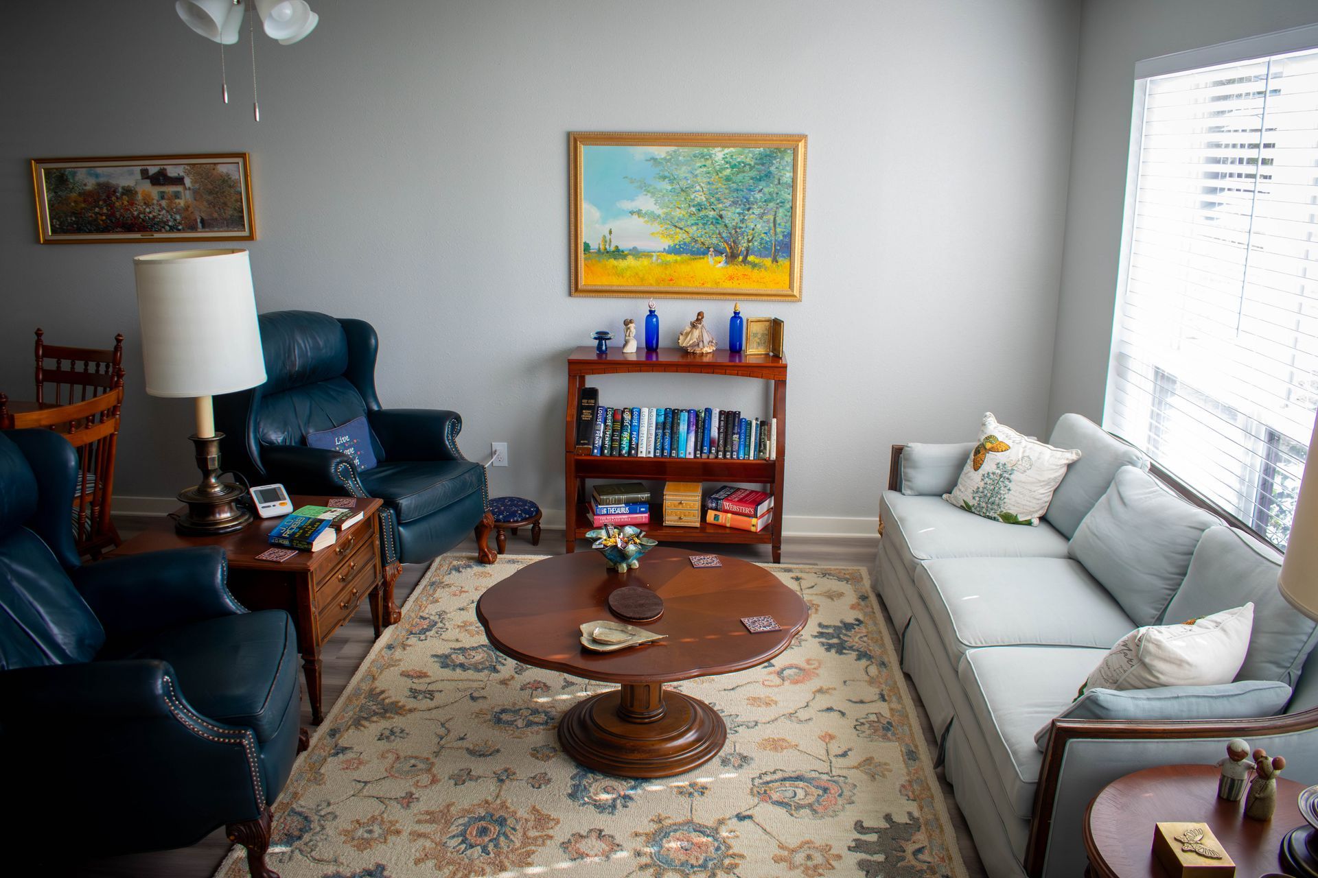 Living room with blue sofa and chairs, rug, round coffee table, bookshelf, and artwork on gray walls.