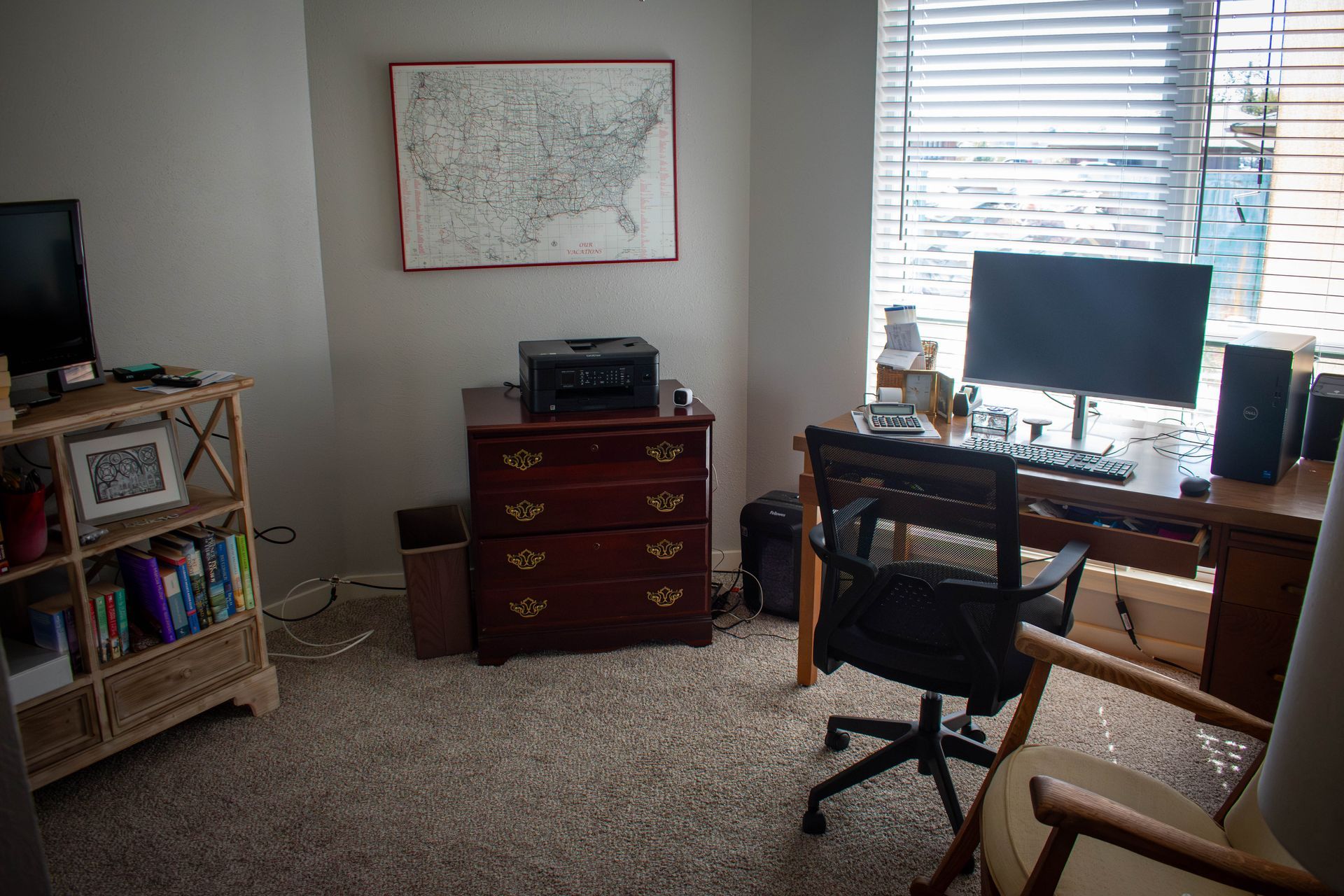 Home office with desk, computer, bookcase, map, and dresser. Neutral tones, carpeted floor, window with blinds.
