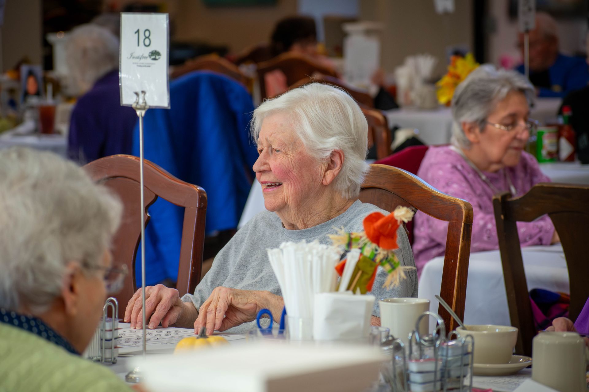 Senior woman smiles at a table in a dining room, others seated around her. Table number 18 visible.