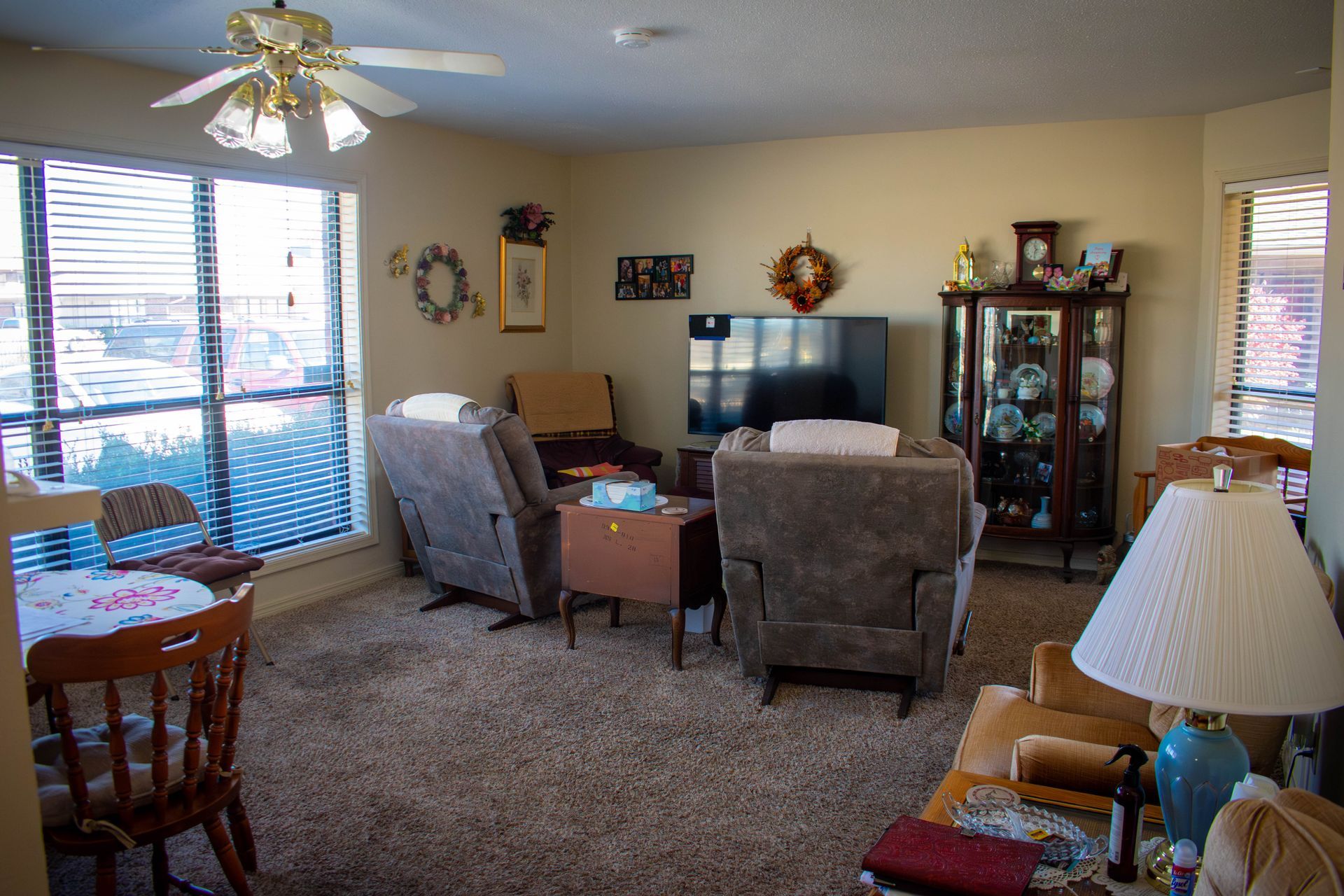 Cozy living room with large windows, recliners, and a dining area. Brown carpet, beige walls, and various decorations.