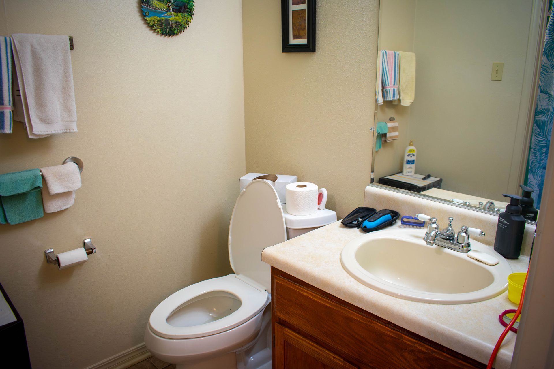 Small bathroom with toilet, sink, mirror, and towel racks. Beige walls, light countertop, and wood cabinet.