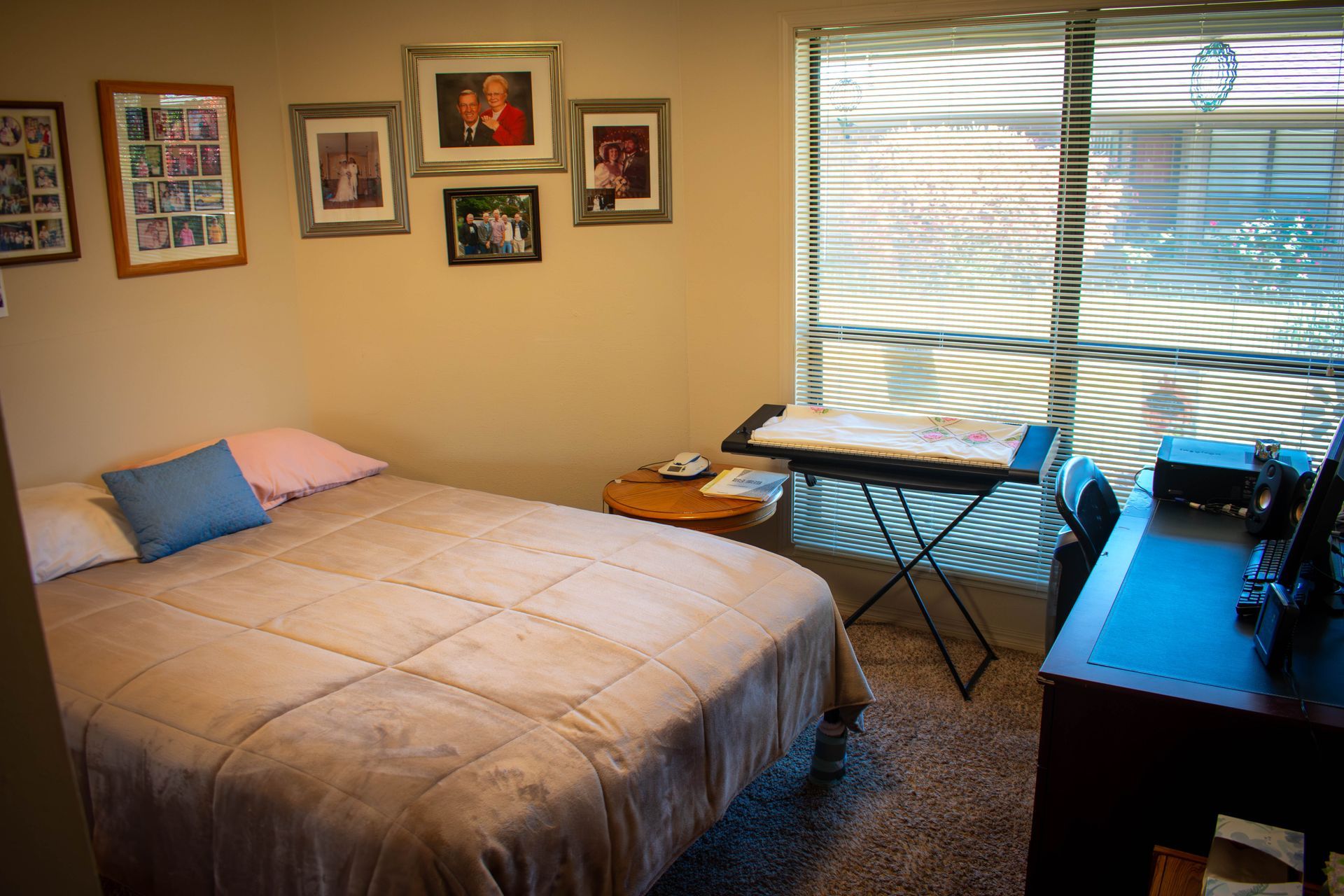 Bedroom with bed, desk, and framed photos on wall. Window with blinds and a drawing table.