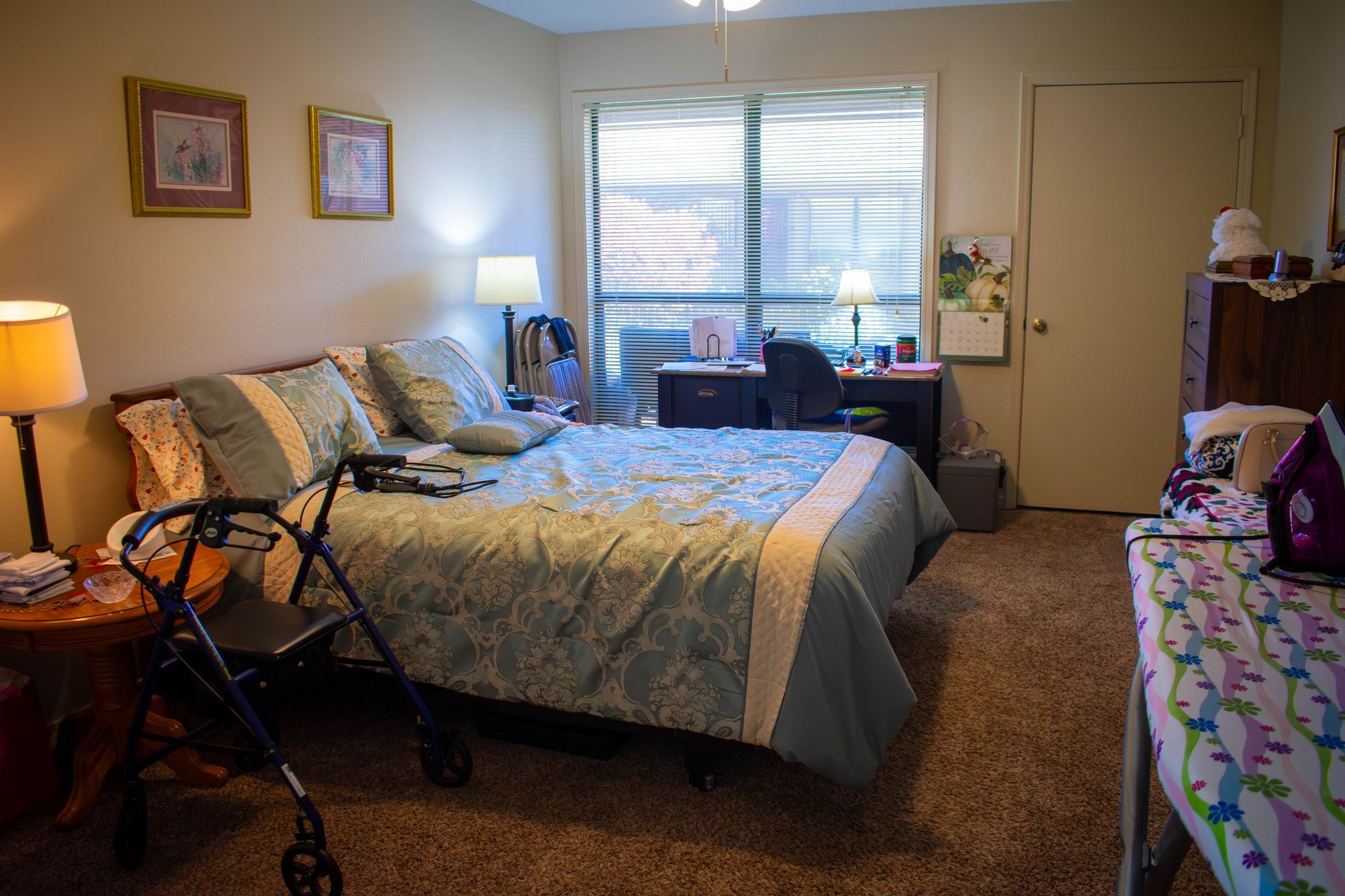Bedroom with bed, desk, walker, and furniture. Window with blinds, pictures, and lamps.