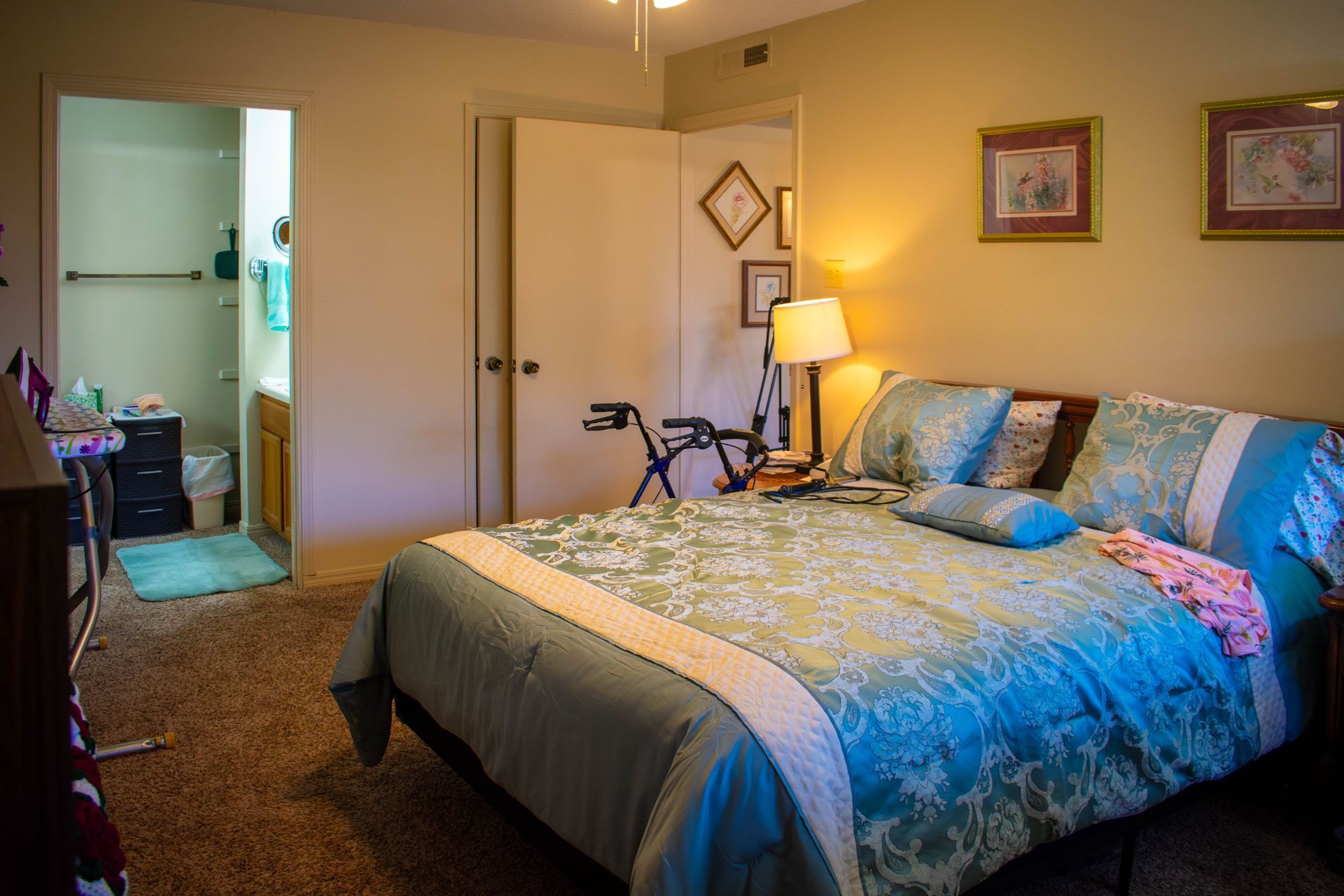 Bedroom with teal bedding, open bathroom door, walker, and framed artwork.