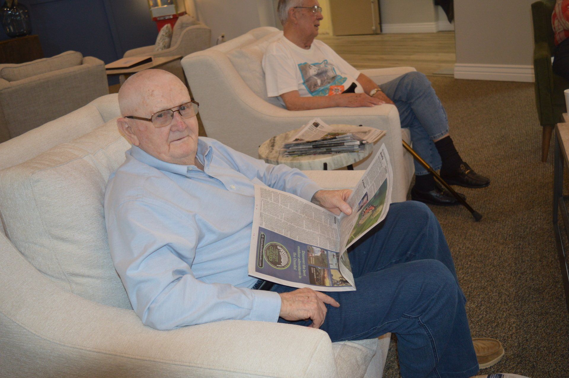 Two older men sitting in armchairs, one reading a newspaper, the other looking on. Beige chairs in a room.