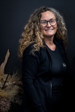 A woman with curly hair and glasses is wearing a black jacket and smiling.