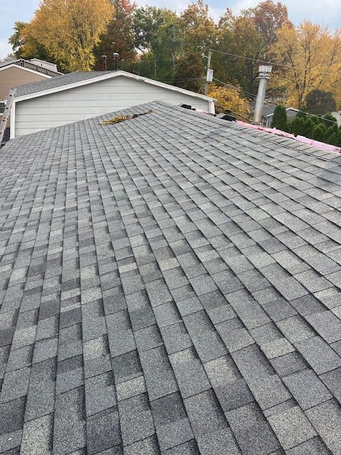 Gray asphalt shingles on a sloped residential roof with a white garage and autumn trees in the background.
