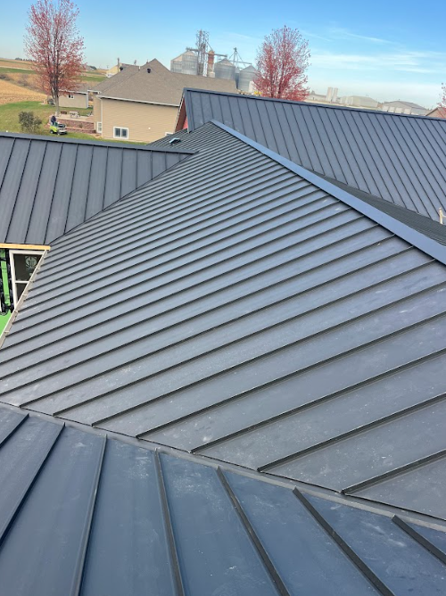 An elevated view of a gray metal roof with standing seams, featuring multiple pitches and a house in the background.