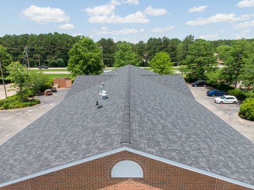 An elevated, outdoor view of a brick building with a dark, shingled roof, surrounded by a parking lot and green trees.