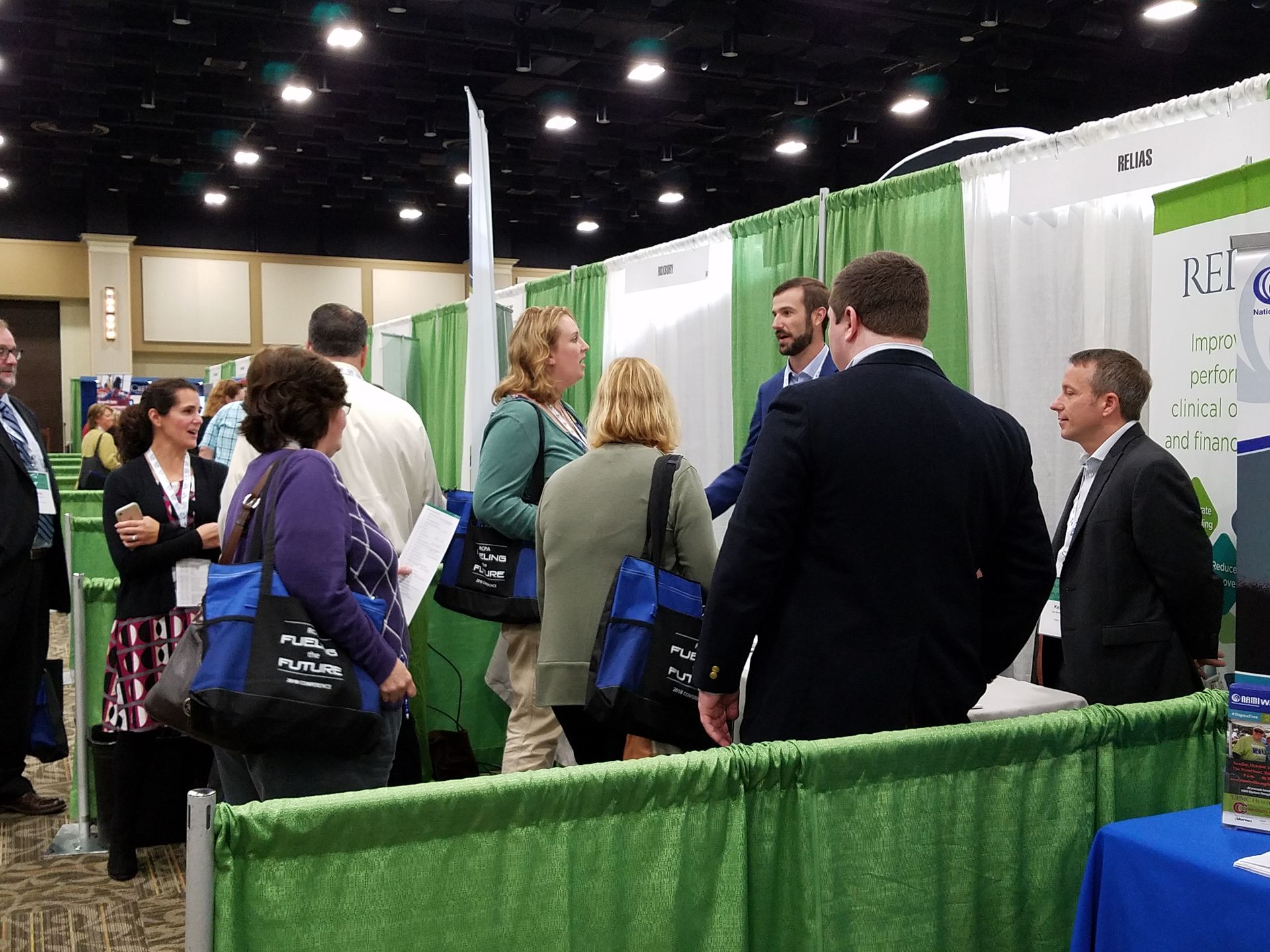 A group of people gathers at a trade show booth, conversing near green curtains under indoor lighting.