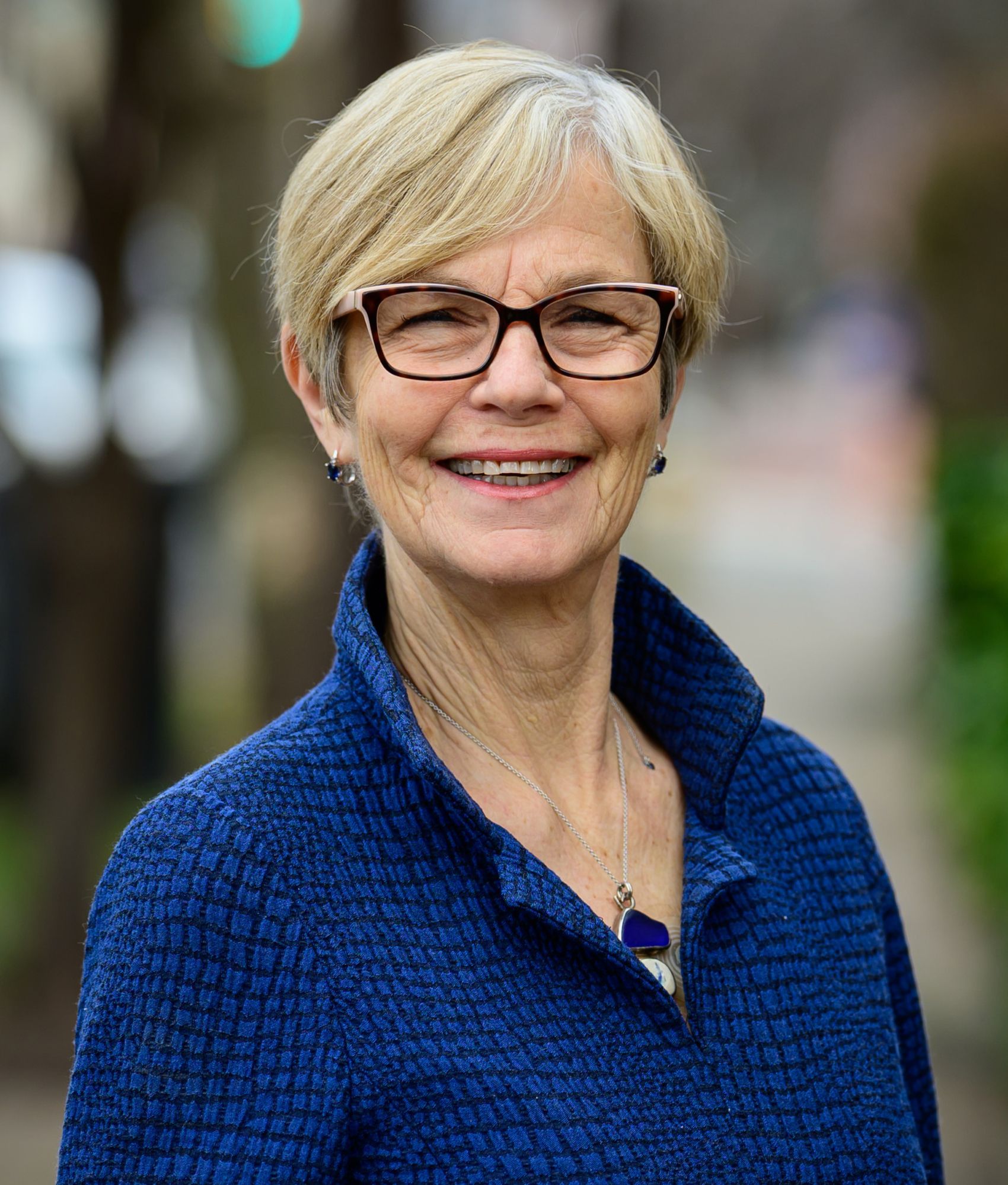 Smiling person in glasses and a blue sweater outdoors with a blurred background
