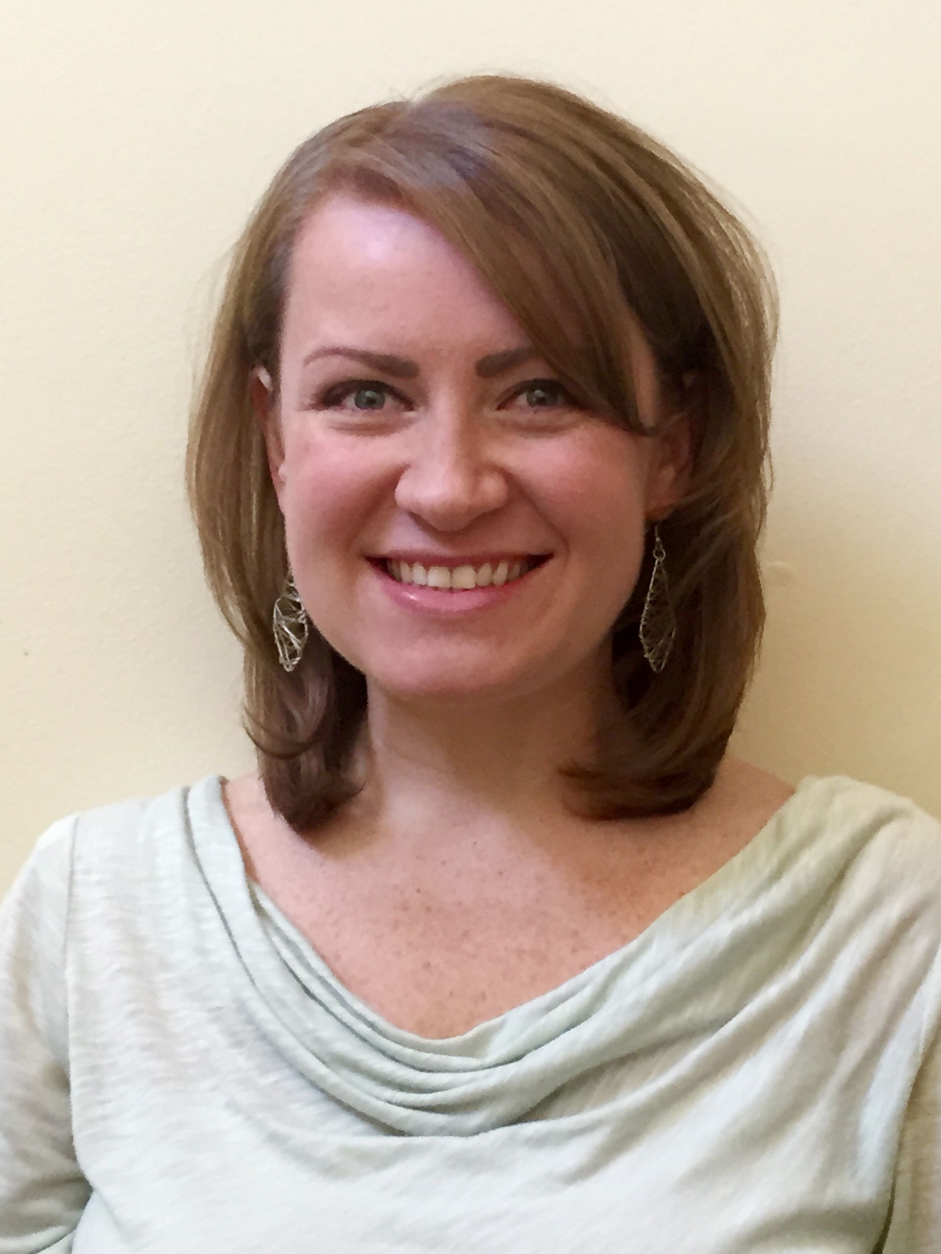 A smiling person with shoulder-length brown hair, wearing a light green cowl-neck top and dangling silver earrings.