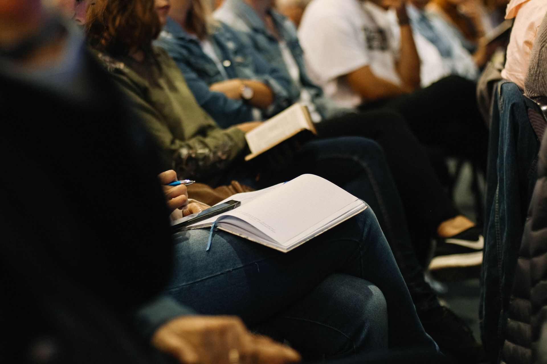 A group of people sitting in rows, some writing in notebooks and reading books during a presentation or workshop.