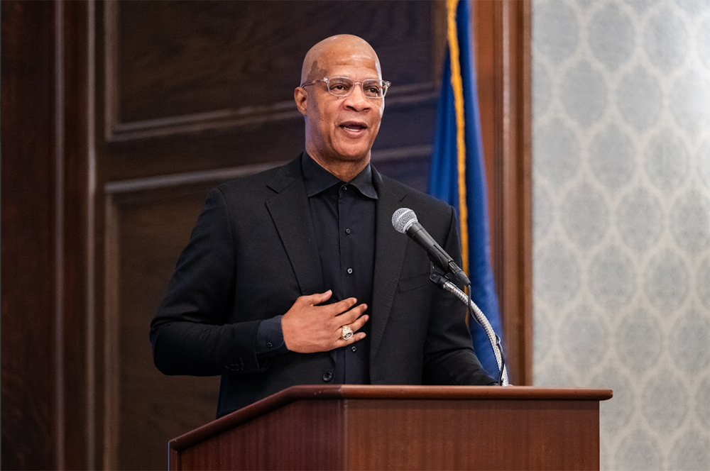 Man speaking at a podium with a microphone in a formal room, hand on chest