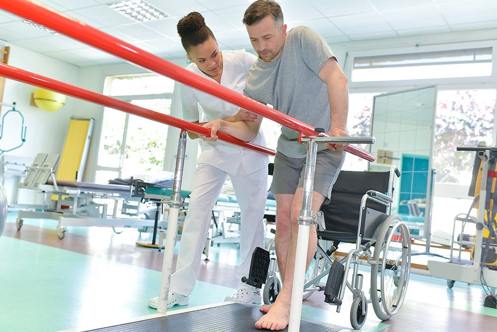Therapist assists man walking with parallel bars in a bright rehab gym; wheelchair nearby