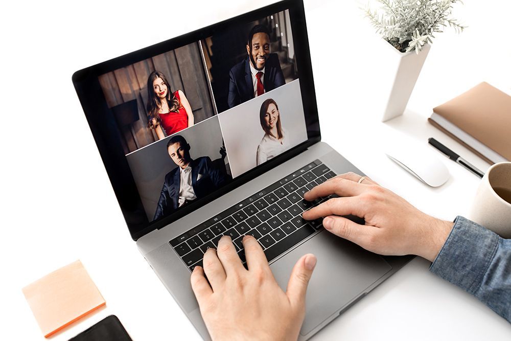 Hands typing on a laptop during a four-person video call on a bright desk