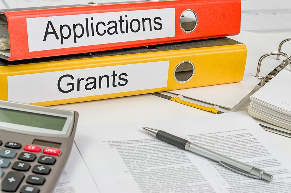 Red binder for Applications on top of a yellow binder for Grants next to office supplies