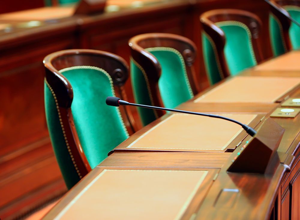 A row of green upholstered chairs with microphones arranged at a wooden desk in a formal meeting room.