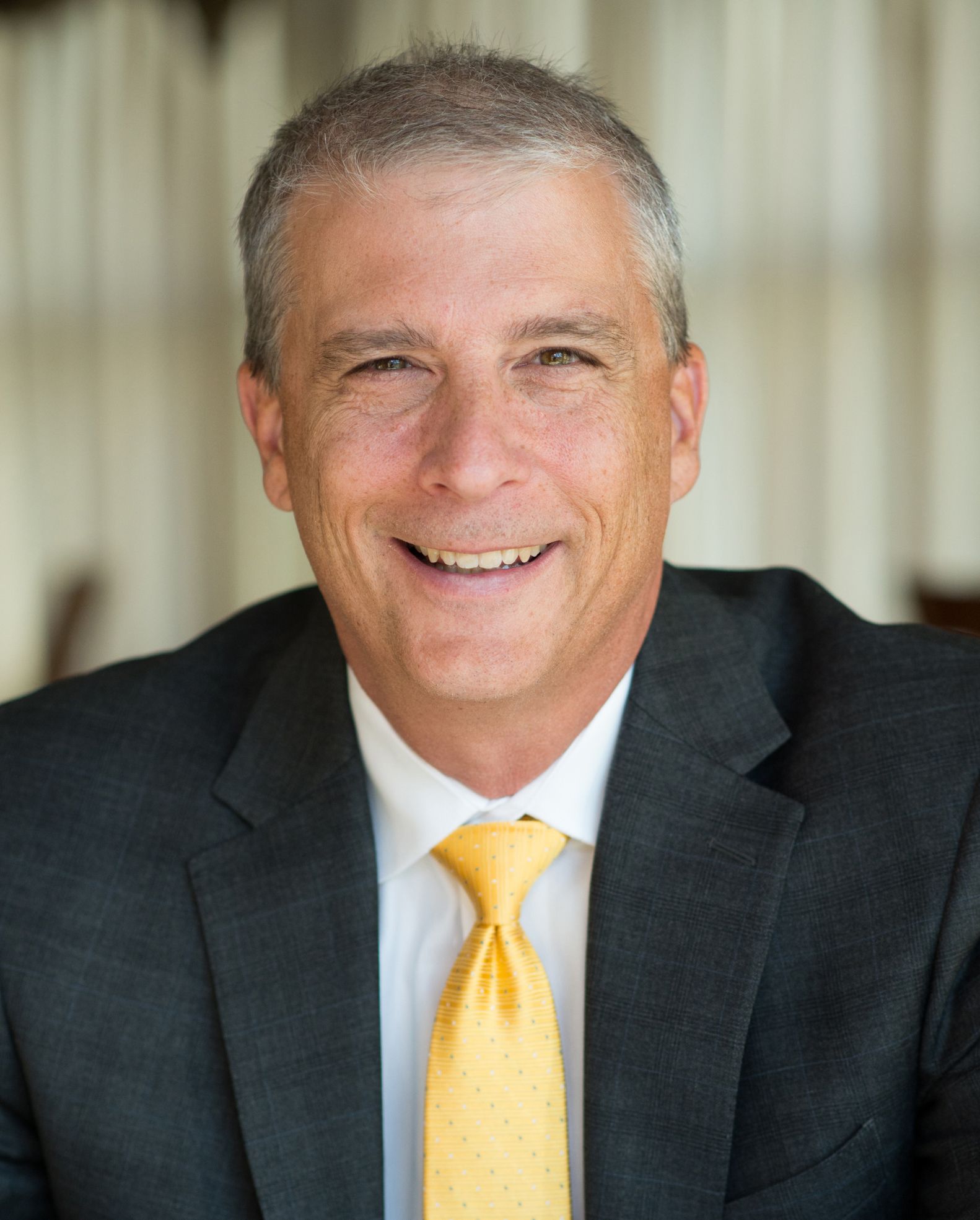 Smiling man in a dark suit and yellow tie, posed indoors against a blurred background.