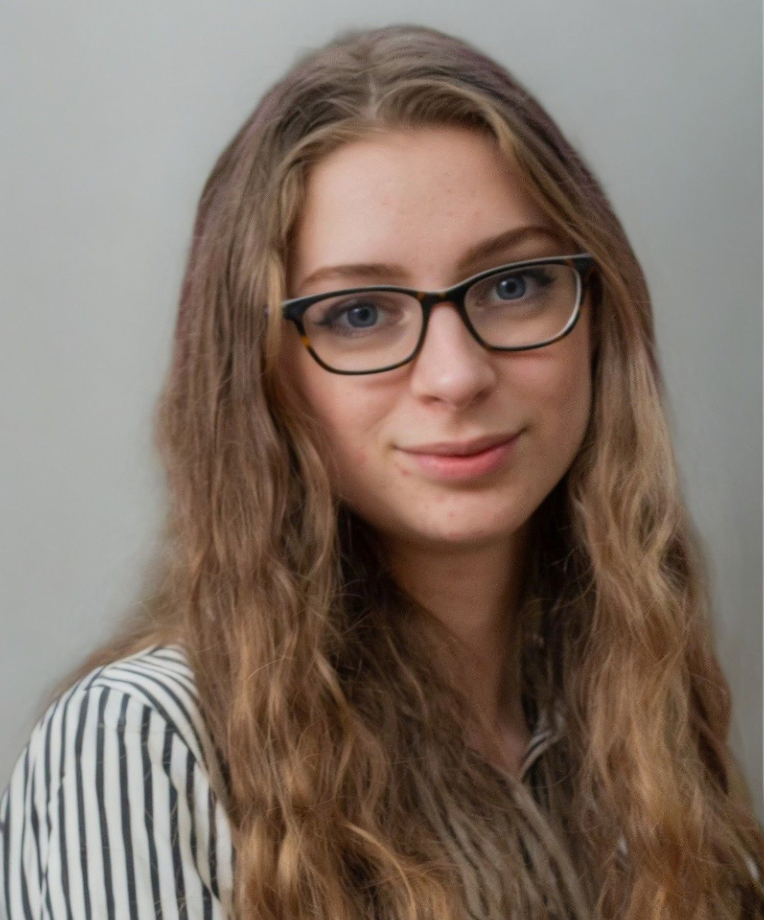 A person with long, wavy brown hair and glasses, wearing a white and black striped shirt against a grey background.