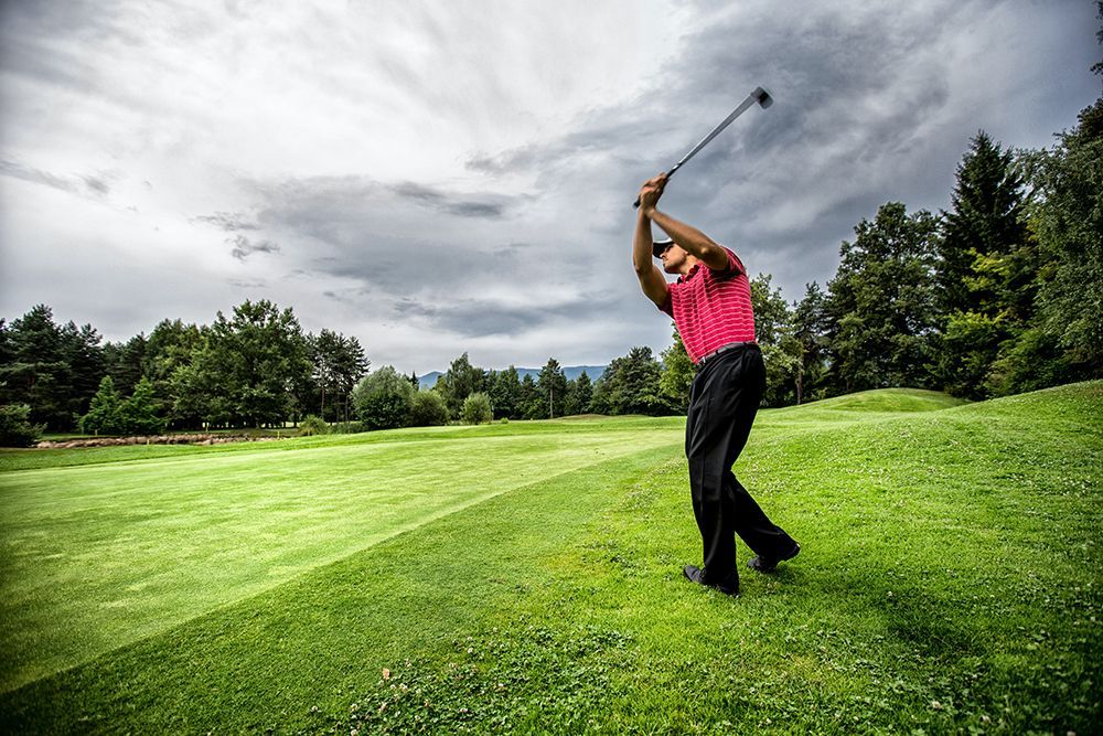 A golfer in a pink shirt swings a club on a grassy course under a cloudy sky.