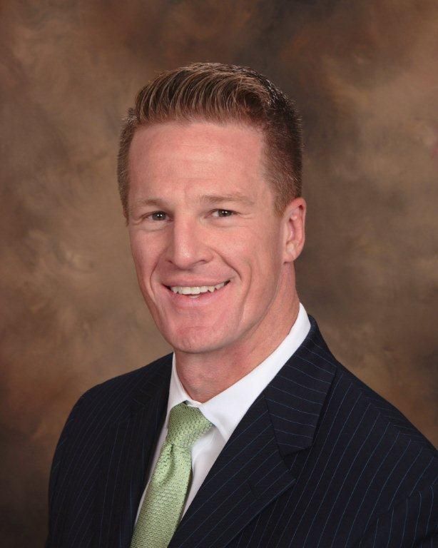 Headshot of a smiling person wearing a dark pinstripe suit, white shirt, and light green tie against a mottled backdrop.